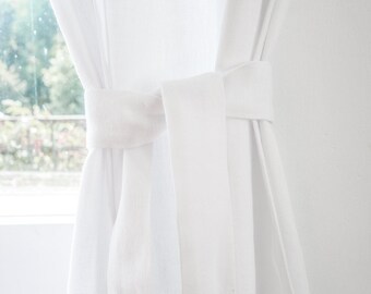 May include: White sheer curtains tied back with a white fabric tie. The curtains are hanging in a window with a view of green trees outside.