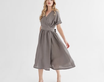 May include: A woman wearing a gray linen wrap dress with short sleeves and a tie at the waist. The dress has a flowing skirt and is knee-length.