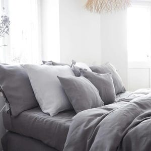 May include: A gray linen duvet cover and pillowcases with a white linen pillow on top. The bedding is arranged on a bed with a white headboard and a window in the background.