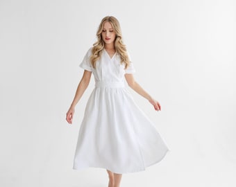Puede incluir: Un vestido blanco de lino con un corpiño de estilo envolvente y una falda fluida. El vestido tiene mangas cortas y un escote en V.