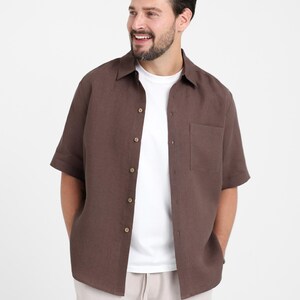 May include: A brown short-sleeved shirt with a button-down front and a chest pocket. The shirt is worn over a white t-shirt and paired with beige pants. The outfit is casual and suitable for warm weather.