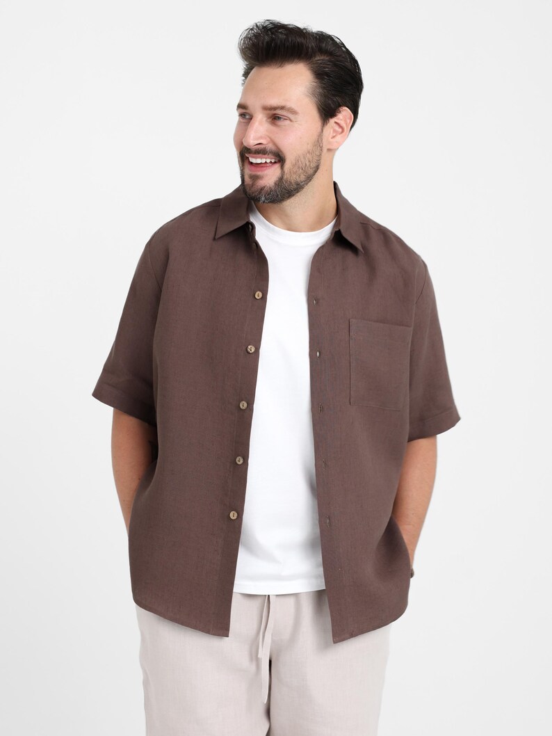 May include: A brown short-sleeved shirt with a button-down front and a chest pocket. The shirt is worn over a white t-shirt and paired with beige pants. The outfit is casual and suitable for warm weather.