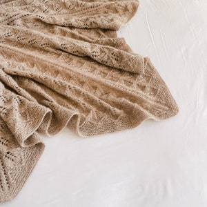 May include: A soft, brown knitted throw blanket with a delicate openwork pattern. The blanket is draped over a white surface.