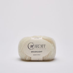Op de afbeelding: Een bol crèmekleurig garen met een label met de tekst "Cardiff Cashmere Brushlight Made in Italy" en een kleine geitenillustratie.