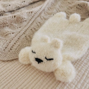 May include: A white knitted plush toy shaped like a bear, lying on a cream-coloured knitted blanket. The bear has black eyes and a black nose.