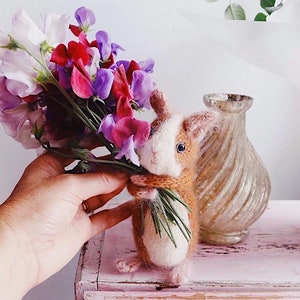 May include: A handmade, brown and white felted bunny figurine holding a bouquet of colorful sweet pea flowers. The bunny has a knitted scarf and is standing on a pink surface. A decorative glass vase is in the background.