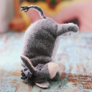 Puede incluir: Un juguete de burro tejido a mano de color gris con una barriga gris claro y un hocico beige. El burro está posicionado boca abajo, con la cabeza tocando la superficie. El burro tiene una cola pequeña y oscura y orejas pequeñas.