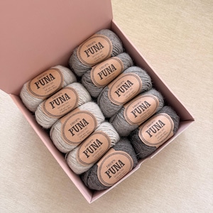 May include: A pink cardboard box containing ten skeins of Drops Puna yarn in various shades of gray. Each skein features a tan paper label with the text Drops Puna Natural Mix, soft and warm, in superfine alpaca, arranged in a grid pattern.