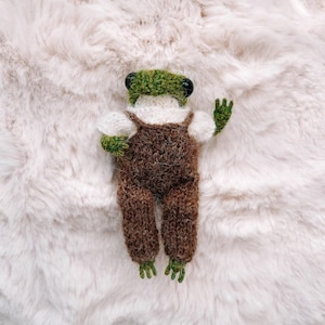 May include: A handmade, knitted frog doll with a green hat and webbed feet. The frog is wearing brown dungarees and white sleeves. The doll is lying on a fluffy, white surface.