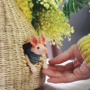 Puede incluir: Un animal de punto rosa y blanco con un suéter a rayas azul y verde asomándose de una cesta tejida. Una mano se extiende hacia el animal.