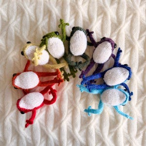 May include: A collection of seven knitted frog toys in a rainbow of colors: red, yellow, green, dark green, purple, blue, and light blue. Each frog has a white belly and black eyes. The frogs are arranged on a white, textured surface.