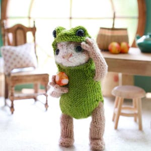 May include: A handmade knitted bunny wearing a green frog costume, holding a small orange and white ball. The bunny is standing, with its right paw raised to its forehead. The background includes a wooden table and chair.