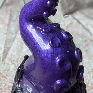 May include: A purple tentacle-shaped decorative object with a glossy finish. The tentacle has multiple suction cups and a curved shape. The base is dark with a textured appearance. The object is likely a decorative item or sculpture.