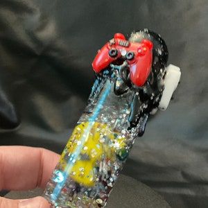 May include: A clear, glitter-filled glass pipe with a yellow and multi-colored design inside. The top of the pipe features a red and black controller design. The pipe is held in a hand.