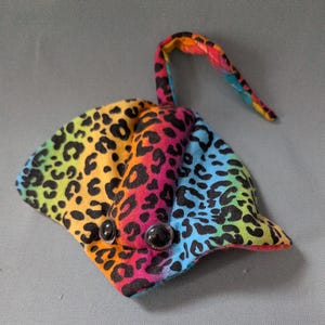 May include: A rainbow-colored stuffed stingray with black spots, resembling a leopard print. The stingray has large black eyes and a long tail.