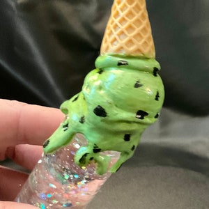 May include: A whimsical, handmade ice cream cone-shaped object. The top features a waffle cone, with a scoop of green ice cream speckled with black dots. The clear base is filled with iridescent glitter.