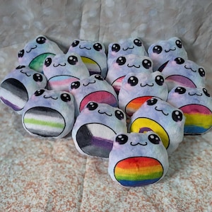 May include: A collection of plush frog toys with various LGBTQ+ pride flag designs. The frogs are light blue with embroidered faces and colorful bellies, including rainbow, pansexual, and asexual flags.