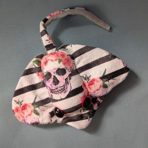 May include: A plush toy shaped like a stingray with a black and white striped body. The body is decorated with pink roses and a pink skull with a floral crown. The stingray has black button eyes.