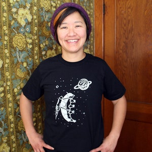 May include: A person wearing a black t-shirt with a white graphic of a creature in space. The creature is holding a rocket and there is a planet in the background. The person is wearing a purple beanie.