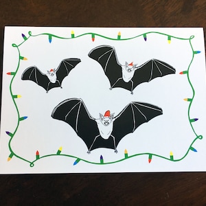 May include: A white card with a green garland of lights around the border. Three black bats with red Santa hats are flying in the center of the card.