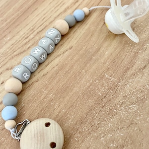 May include: A wooden and silicone baby teether with a clip and a dummy clip. The teether has beads with the name "ROWAN" spelled out on them. The beads are in shades of grey and beige.