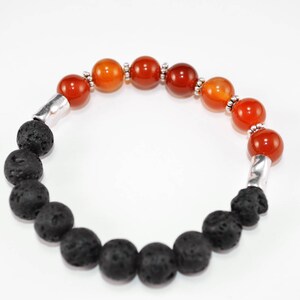 Puede incluir: Un brazalete hecho con cuentas de lava negra y cuentas de cornalina naranja. El brazalete tiene detalles metálicos plateados.