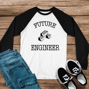 Puede incluir: Camiseta blanca de manga larga con mangas negras. La camiseta tiene un gráfico en blanco y negro de bloques de construcción y el texto "FUTURE ENGINEER".