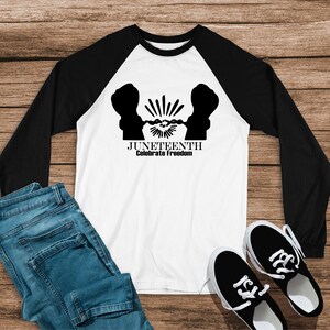 Puede incluir: Camiseta de manga larga de estilo béisbol en blanco y negro con un gráfico en blanco y negro de dos figuras rompiendo cadenas. El texto "Juneteenth Celebrate Freedom" está impreso debajo del gráfico.