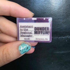 May include: A purple and white plastic name tag with the text "Assistant to the Regional Manager, Sales, Dunder Mifflin Paper Company, Scranton, PA".