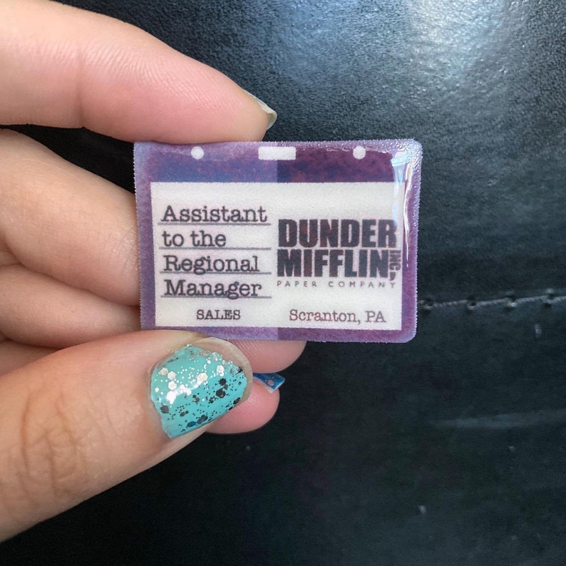 May include: A purple and white plastic name tag with the text "Assistant to the Regional Manager, Sales, Dunder Mifflin Paper Company, Scranton, PA".