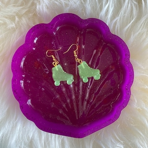 May include: A pair of green roller skate earrings with gold hooks. The earrings are displayed in a purple shell-shaped dish.