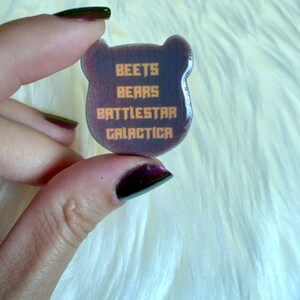 May include: A purple bear-shaped resin pin with the text "BEETS BEARS BATTLESTAR GALACTICA" in orange.