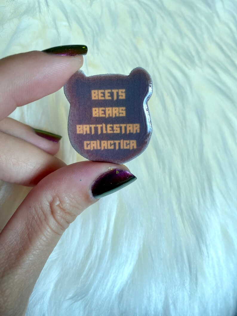May include: A purple bear-shaped resin pin with the text "BEETS BEARS BATTLESTAR GALACTICA" in orange.