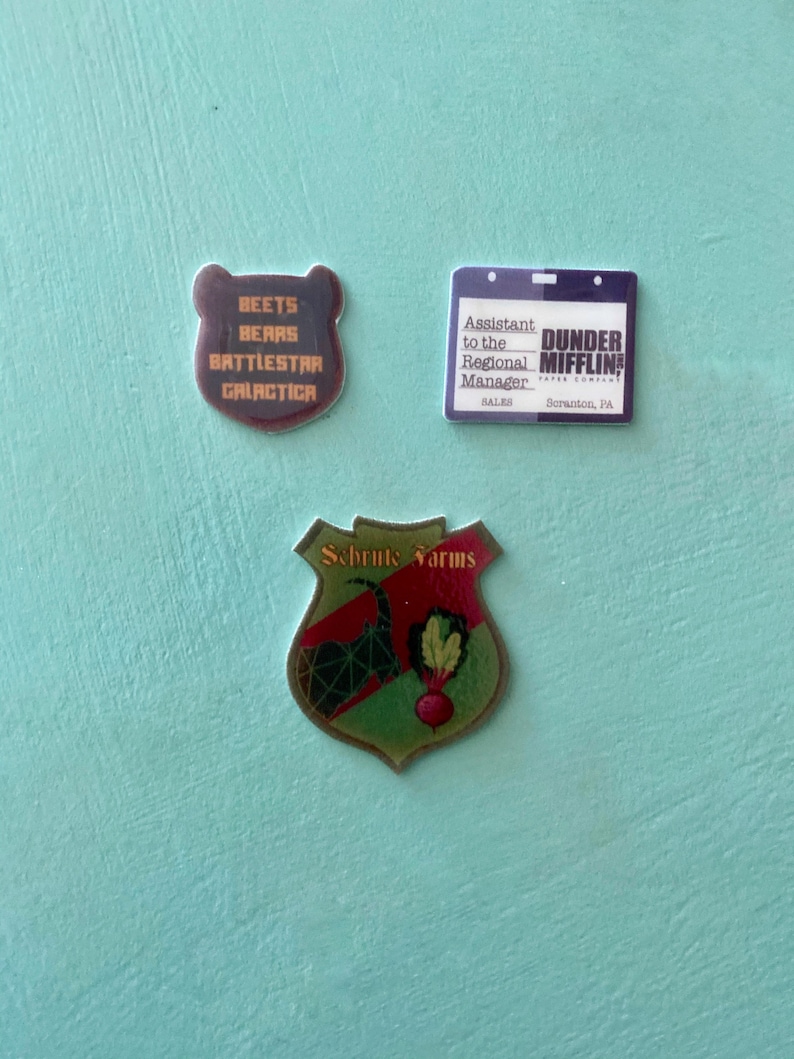 May include: Three stickers featuring characters and text from the TV show The Office. The first sticker is a brown bear with the text "Beets Bears Battlestar Galactica". The second sticker is a blue and white ID badge with the text "Assistant to the Regional Manager Sales Dunder Mifflin Paper Company Scranton, PA". The third sticker is a green and gold shield with the text "Schrute Farms" and a red beet and green leaves.