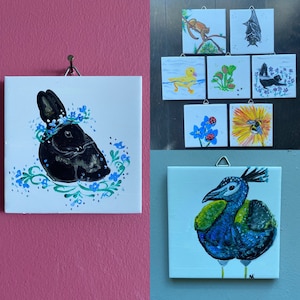 May include: A set of 10 hand-painted square tiles featuring various animal and nature scenes. The tiles are white with black outlines and colorful details. The scenes include a rabbit with blue flowers, a duck, a bee, a bat, a squirrel, and a peacock.