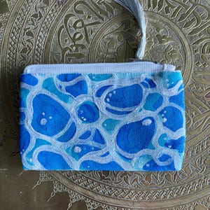 May include: A small, rectangular coin purse with a blue and white abstract pattern. The purse has a white zipper and a light blue tassel attached to the zipper pull. The pattern features organic shapes in shades of blue.