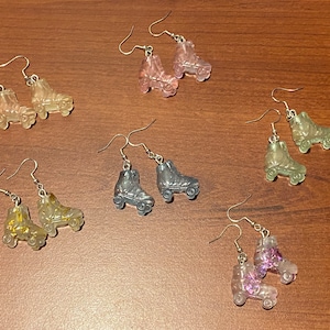 May include: A set of ten pairs of roller skate earrings. The earrings are made of clear resin and have a variety of colors, including pink, purple, blue, and green. Some of the earrings have glitter embedded in the resin.