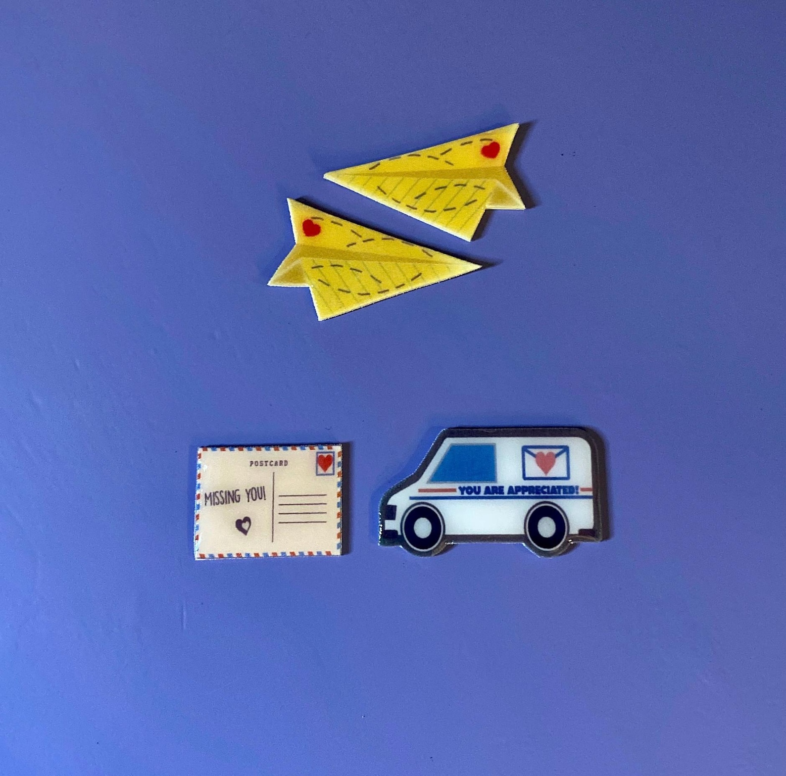 In clockwise order on a blue background, 3 pins; 2 yellow lined paper style folded paper airplanes with matching red hearts and drawn lines to indicate movement of the hearts, a mail truck with an emblem of an envelope sealed with a heart and the words "YOU ARE APPRECIATED", and a post-card style pin with a heart in the place of the stamp, the words "POSTCARDS" as well as "MISSING YOU" and a heart.