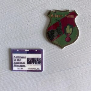 May include: A purple and white plastic name tag with the text "Assistant to the Regional Manager" and "DUNDER MIFFLIN PAPER COMPANY" and "SALES Scranton, PA".  A green and red shield with the text "Schrute Farms" and a black and green image of a beet.