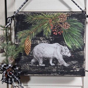 May include: A black wooden sign with a white polar bear walking through a pine forest. The sign has a distressed look and is decorated with pine branches, pine cones, and red berries.