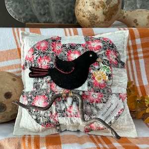 May include: A decorative pillow with a blackbird applique perched on a twig. The pillow features a patchwork design with red floral fabric and a white background. The blackbird has red accents and holds a flower.