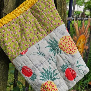 May include: Quilted oven mitt with a yellow border and a floral pattern on the top half. The bottom half features a pineapple design with red and yellow fruit and green leaves. The mitt is hanging on a wooden fence.