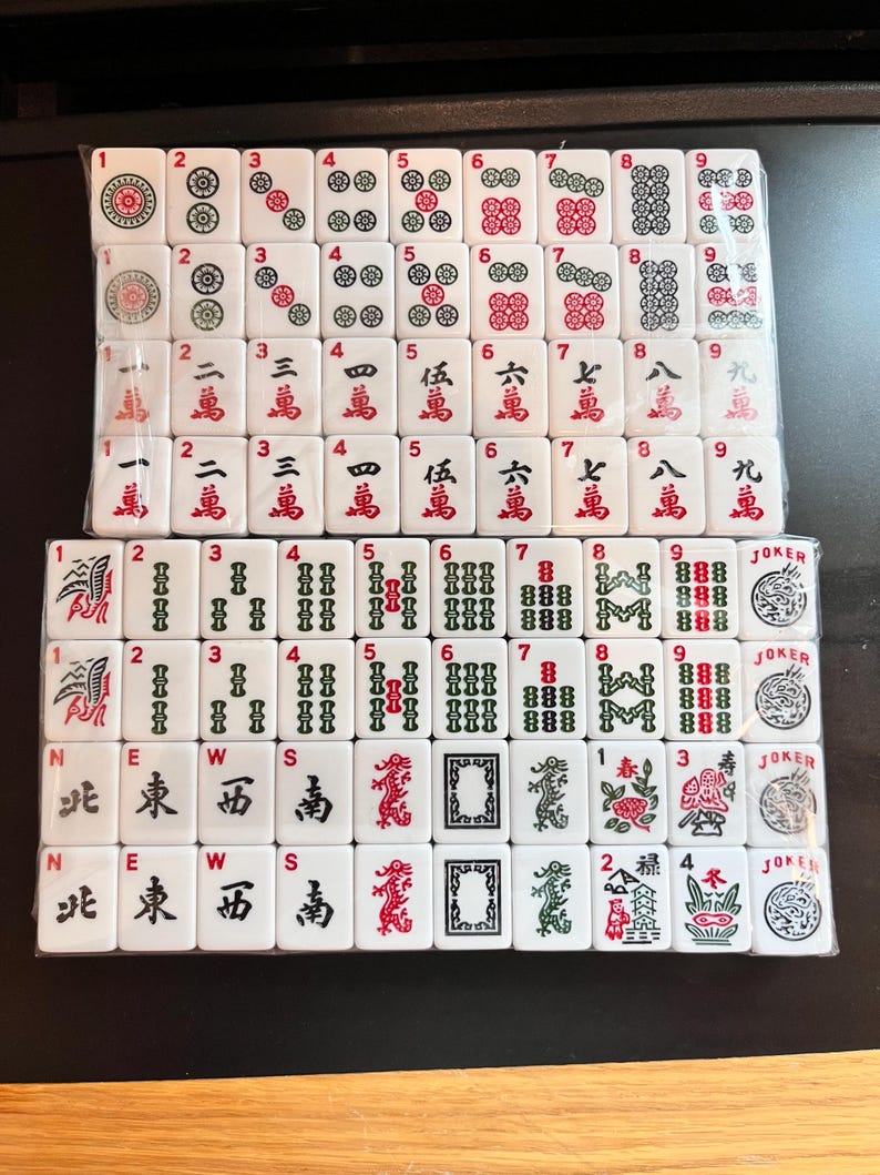 May include: A complete set of white Mahjong tiles, featuring various symbols, numbers, and Chinese characters in red and black. The tiles are arranged in rows, showcasing the different suits and special tiles, including Jokers.