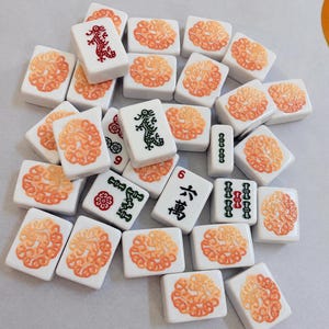 Op de afbeelding: Een verzameling witte rechthoekige mahjongtegels met oranje cirkelvormige bloempatronen op de achterkant. Sommige tegels tonen groene of rode draaksymbolen, numerieke tekens en kleine bloempictogrammen in zwarte, rode en groene inkt.