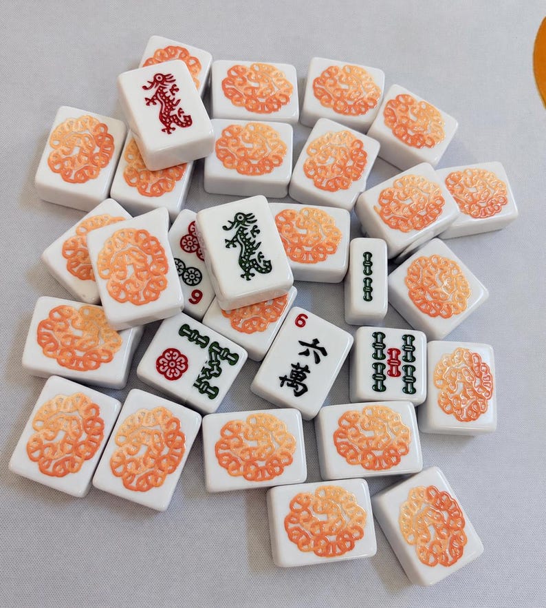 Op de afbeelding: Een verzameling witte rechthoekige mahjongtegels met oranje cirkelvormige bloempatronen op de achterkant. Sommige tegels tonen groene of rode draaksymbolen, numerieke tekens en kleine bloempictogrammen in zwarte, rode en groene inkt.