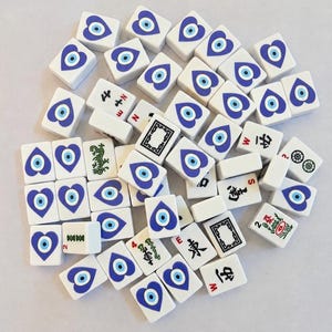 May include: A collection of white rectangular mahjong tiles featuring a blue and white heart-shaped evil eye design on the faces. Several tiles display black Chinese characters, green bamboo patterns, and red directional letters like N, S, E, and W.