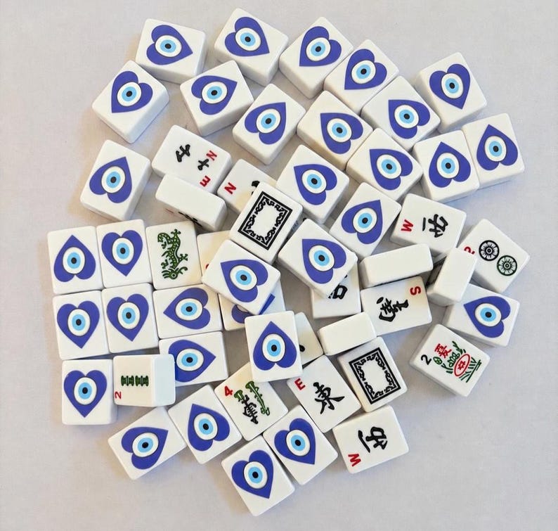 May include: A collection of white rectangular mahjong tiles featuring a blue and white heart-shaped evil eye design on the faces. Several tiles display black Chinese characters, green bamboo patterns, and red directional letters like N, S, E, and W.