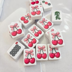 May include: A collection of white rectangular mahjong tiles scattered on a surface. Most tiles feature a graphic of two red cherries tied with a pink ribbon. Other tiles display a green dragon, a black floral pattern with the number 8, or circles.