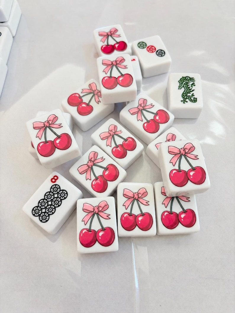 May include: A collection of white rectangular mahjong tiles scattered on a surface. Most tiles feature a graphic of two red cherries tied with a pink ribbon. Other tiles display a green dragon, a black floral pattern with the number 8, or circles.