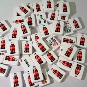 May include: A collection of white rectangular mahjong tiles featuring a Coca-Cola bottle graphic on the front. Some tiles display the red Coca-Cola logo, while others show green numerical symbols or a black and white graphic of a dragon. The tiles are scattered.
